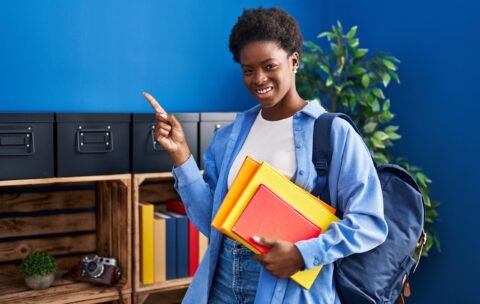 African american woman wearing student backpack and holding books smiling happy pointing with hand and finger to the side